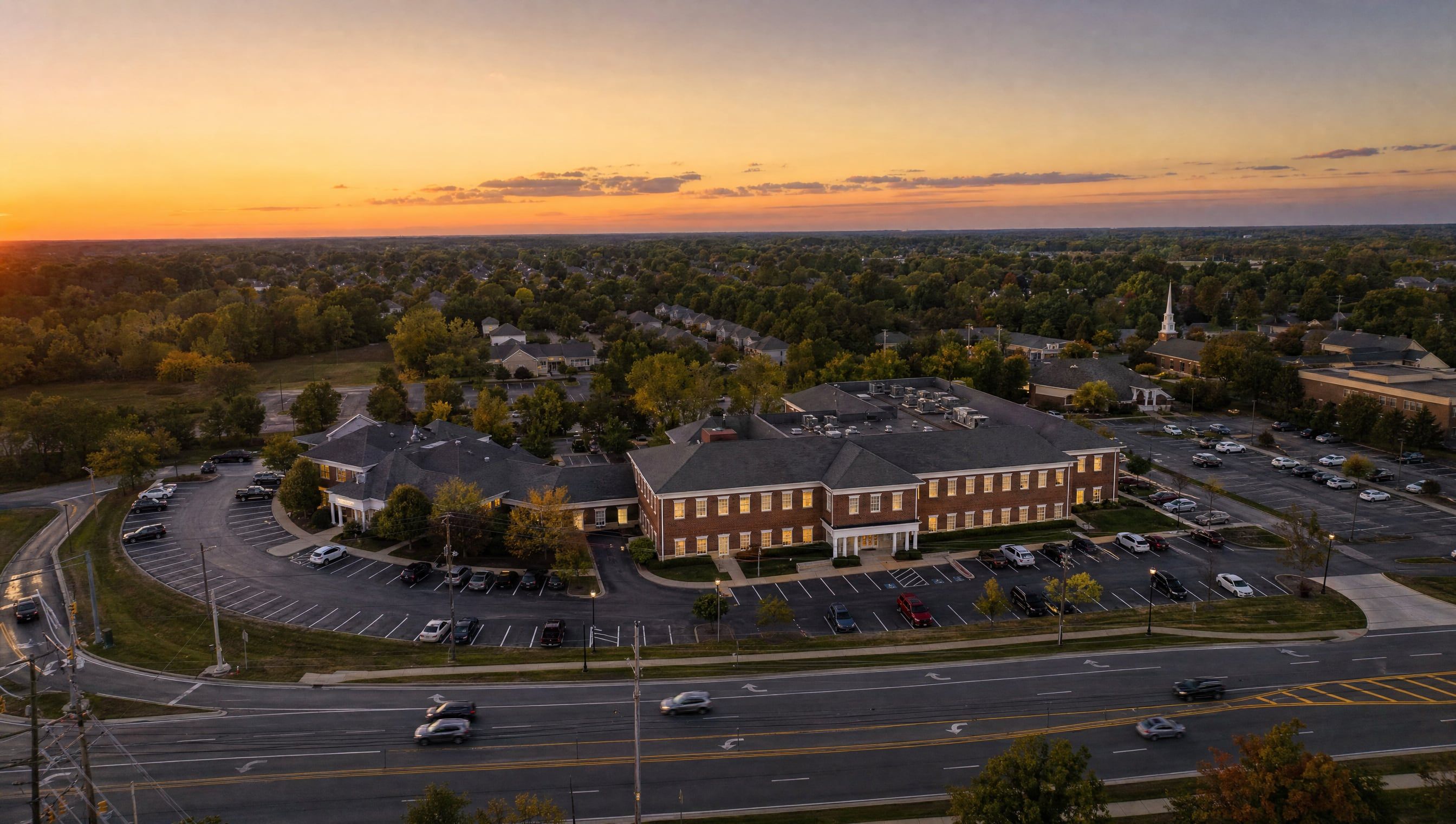 Aerial rendering at sunset of the brick office building at 3985 Medina Road, set back from Route 18 just east of Foote Road, with trees and a church steeple beyond